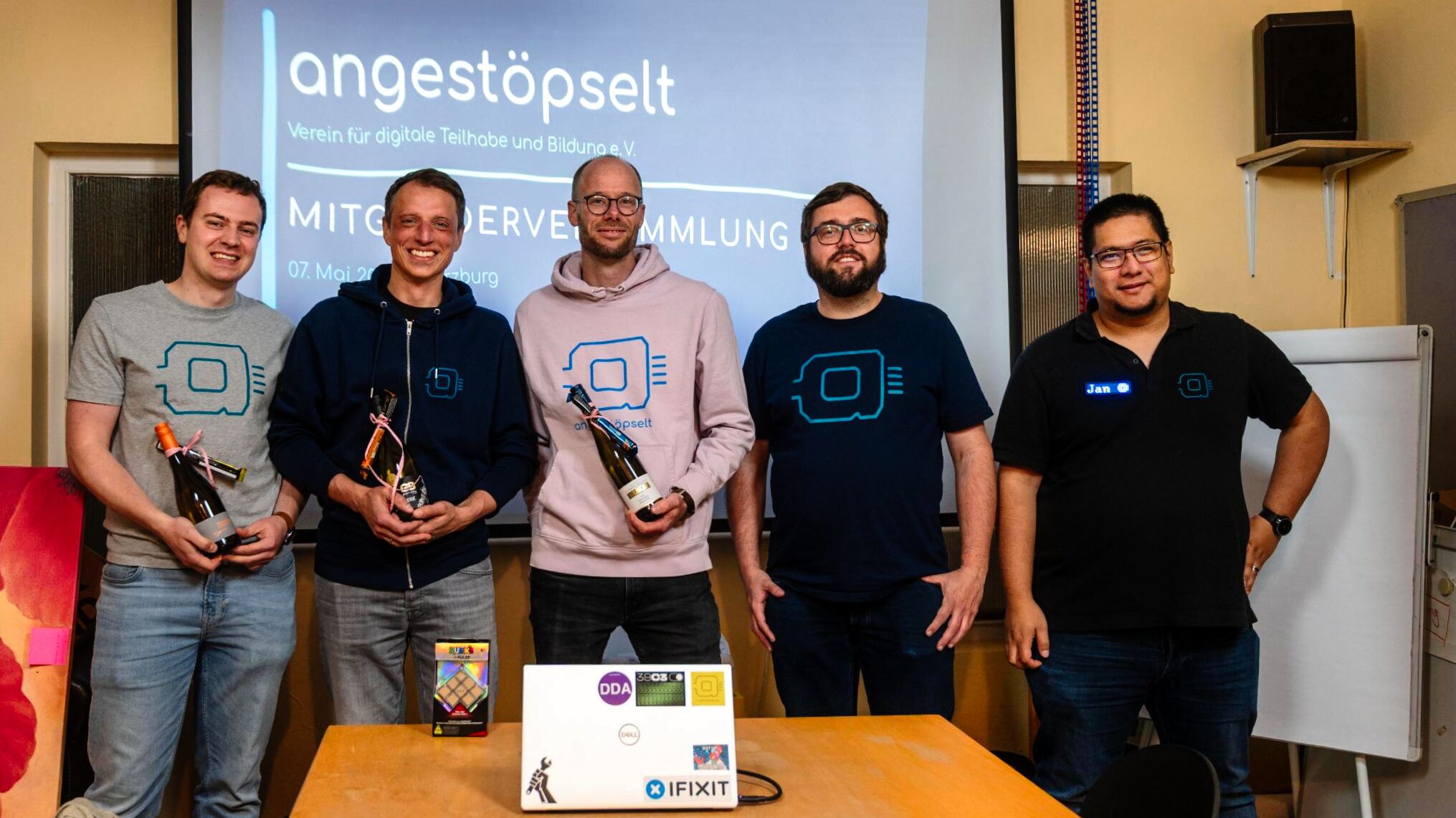 Fünf Personen blicken in die Kamera und stehen vor einem Projektor-Schriftzug: angestöpselt Mitgliederversammlung. Drei von ihnen (von links) halten kleine Präsente in der Hand.